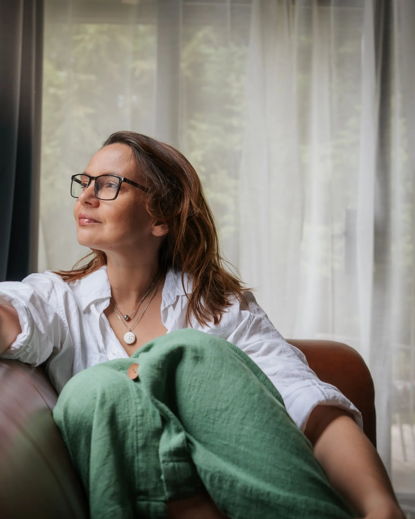 A woman sits in an armchair beside a bright window, looking out.